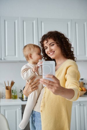 joyful woman taking selfie on mobile phone with toddler daughter holding baby bottle in kitchen