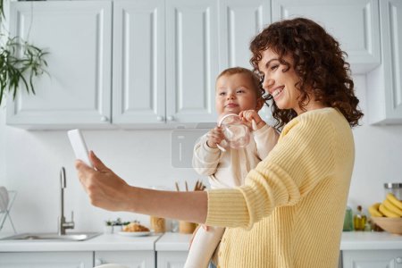 madre emocionada tomando selfie en el teléfono inteligente con niño pequeño sosteniendo biberón, momento sincero