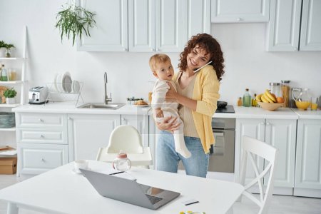 Frau hält Baby in der Hand und spricht auf Smartphone neben Laptop in moderner Küche, Work-Life-Balance