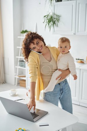 happy woman using laptop while holding child and talking on smartphone in kitchen, multitasking