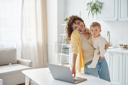 busy mother talking on smartphone and using laptop while holding toddler daughter in kitchen