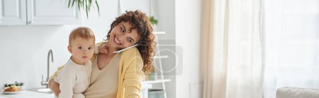freelance work, smiling woman holding little kid while talking on smartphone in kitchen, banner