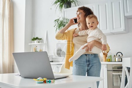 carrera y paternidad, mujer feliz hablando en el teléfono inteligente y sosteniendo el bebé cerca del ordenador portátil en la cocina