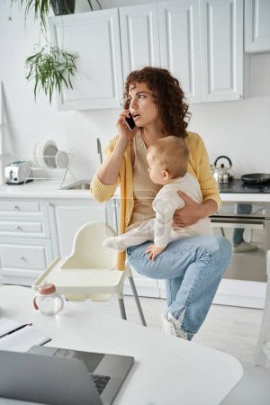 serious woman talking on smartphone and holding little girl near laptop in kitchen, busy mother