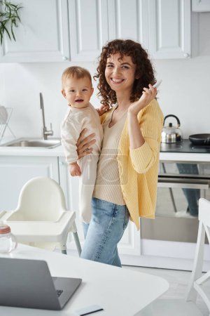 freelancer femenina con smartphone y niño pequeño en las manos sonriendo a la cámara cerca del portátil en la cocina