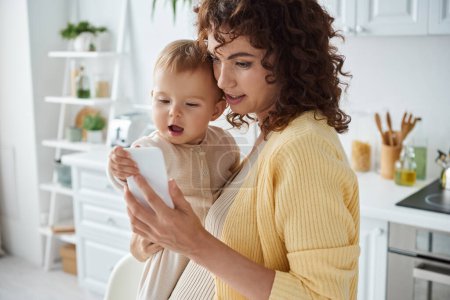 woman holding excited child and browsing internet on smartphone in kitchen, modern parenting