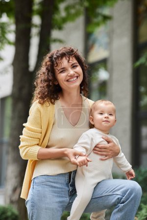 portrait of smiling and curly woman with cute toddler daughter on urban street, happy motherhood