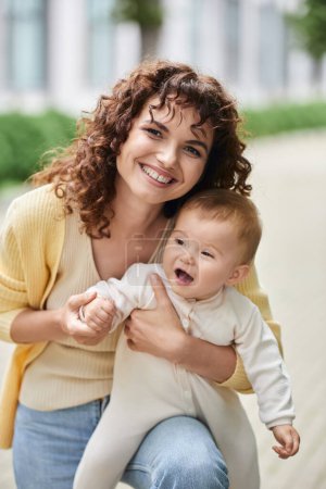 happy woman with wavy hair smiling at camera near little daughter outdoors, family portrait