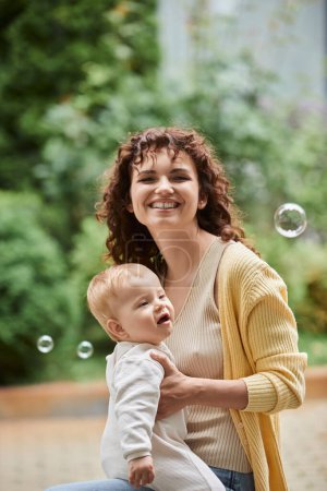 Foto de Retrato de mujer excitada con bebé hija cerca de burbujas de jabón volador al aire libre, momentos felices - Imagen libre de derechos
