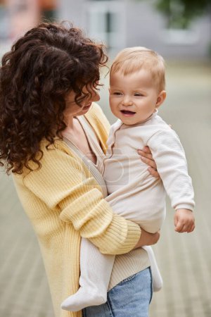 woman with wavy hair standing outdoors and holding adorable toddler daughter in hands, quality time
