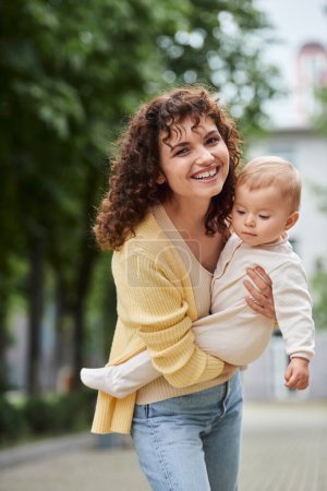 curly woman smiling at camera while holding little daughter on urban street, happy modern family