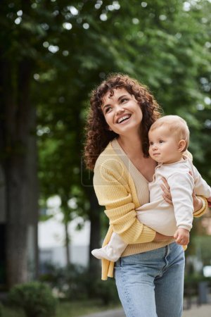 joyful curly woman holding toddler daughter on city street and looking away, bonding moments