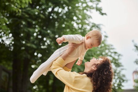 Foto de Mujer emocionada criando a su hijita en las manos mientras se divierte al aire libre, maternidad feliz - Imagen libre de derechos