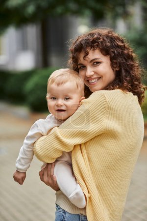 overjoyed woman with wavy hair embracing toddler daughter and smiling at camera outdoors, happiness