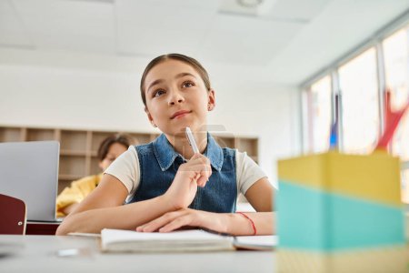 A young girl sitting at a desk, deep in thought, holds a pen, surrounded by a vibrant classroom