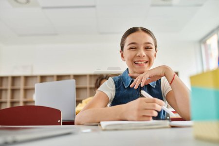 Ein fröhliches junges Mädchen sitzt an einem Schreibtisch und lächelt strahlend, während sie sich auf ihre Umgebung einlässt..