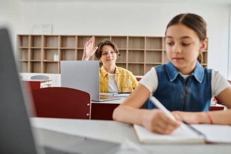 Un jeune garçon assis à un bureau dans une salle de classe lumineuse, concentré sur l'ordinateur portable, engagé dans des études aux côtés de ses pairs.