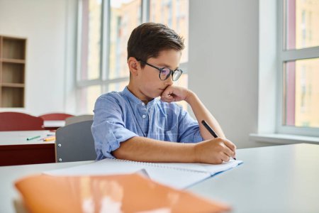 A young boy sits at his desk in a bright classroom, focused on writing on a piece of paper while the teacher instructs the diverse group of kids.