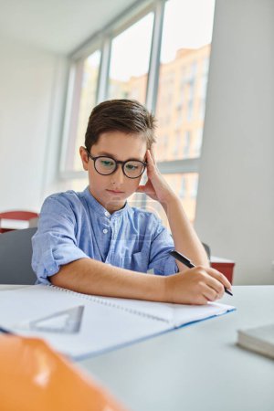 Ein kleiner Junge mit einem Stift in der Hand, konzentriert und engagiert, sitzt an einem Schreibtisch mit Papier in einem lebhaften Klassenzimmer.