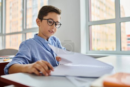 A young boy with glasses sits at a desk, surrounded by papers as he studies intently.
