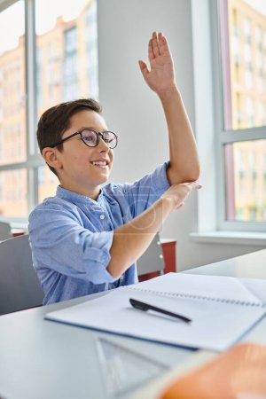 Un niño se sienta en un escritorio en un salón de clases luminoso y animado, levantando la mano para participar en la lección.