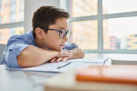 Un jeune garçon concentré portant des lunettes assis avec attention à un bureau, absorbé par l'étude ou le travail sur une tâche.