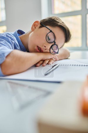 boy wearing glasses peacefully sleeps on a desk in a bright, lively classroom setting