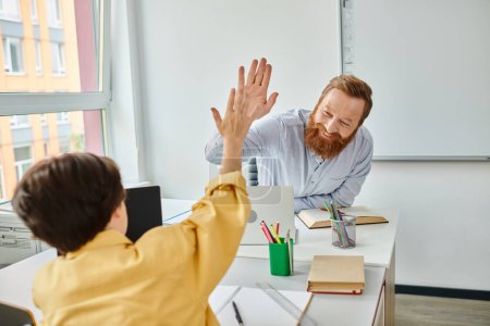 A boy in a yellow shirt is giving a high five to a teacher in a bright, lively classroom setting
