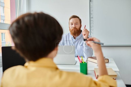 Un homme barbu est assis à un bureau, engagé dans la pensée, peut-être préparer une leçon ou faire des recherches.