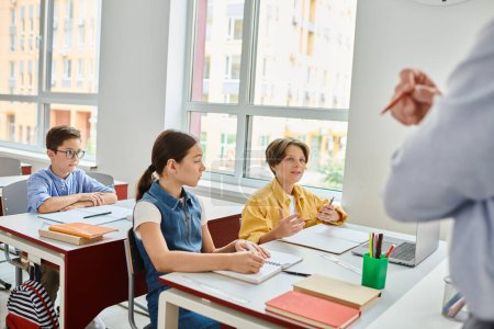 Un groupe d'enfants, absorbés par l'apprentissage, s'assoient avec attention aux bureaux tandis qu'un enseignant masculin vivant transmet des connaissances dans un cadre de classe lumineux.