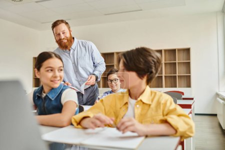 Un grupo de niños se sientan atentamente en los escritorios de una clase iluminada, absortos en la lección de computación que les enseña su maestro masculino.
