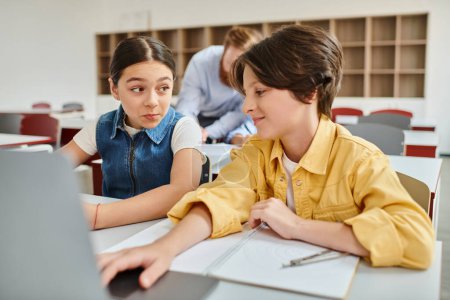 A boy and a girl engage attentively at a table with a laptop, absorbed in a shared learning experience