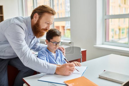Ein Mann sitzt an einem Schreibtisch und unterrichtet einen kleinen Jungen in einem lebhaften Klassenzimmer.
