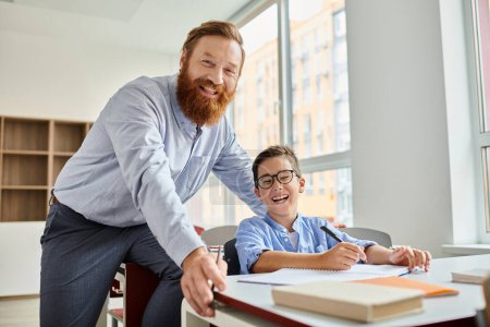 Un hombre de pie junto a un niño en un escritorio, dedicado a una actividad de aprendizaje en un ambiente vibrante en el aula.