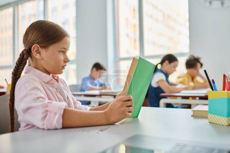 A young girl sits at a table, engrossed in a book in a bright classroom setting.