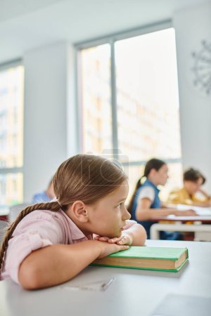 Une jeune fille avec des nattes assise à un bureau, complètement absorbée par la lecture d'un livre dans une salle de classe animée.