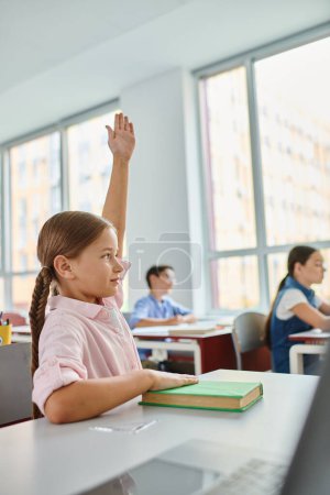 A young girl with long hair raises her hand in a colorful, lively classroom.