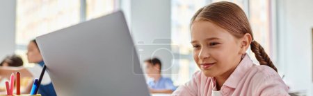 A young girl, sitting in front of a laptop computer in a bright classroom, is engaged in exploring the virtual world