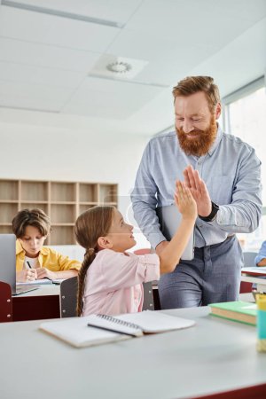 Un homme se tient à côté d'une petite fille dans une salle de classe dynamique, s'engageant dans une activité d'apprentissage avec un groupe d'enfants.