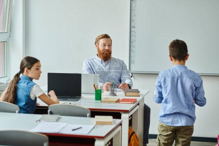 Un hombre se sienta en un escritorio frente a un grupo de niños, involucrándolos en un ambiente animado en el aula.