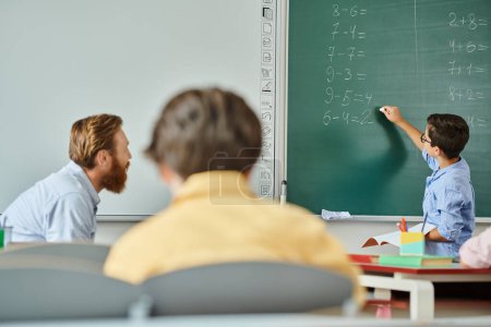 A male teacher instructs a group of students at a table in front of a blackboard in a bright, lively classroom setting.