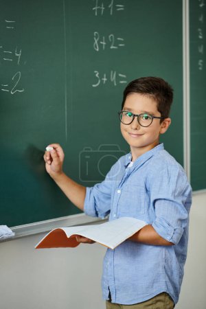 Ein kleiner Junge steht selbstbewusst vor einer Tafel und lernt in einem lebhaften Klassenzimmer.