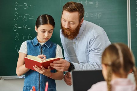 A man stands next to a little girl in front of a blackboard, engaged in a lesson in a bright, lively classroom setting.