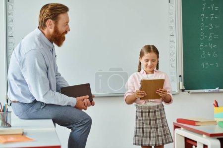 A man, possibly a teacher, sits on a chair next to a little girl, engaging in a moment of mentorship or guidance.