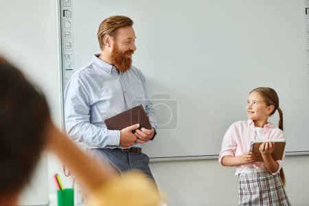 Foto de Un hombre está al lado de una niña frente a una pizarra en un salón de clases brillante, participando en el aprendizaje interactivo. - Imagen libre de derechos