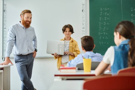 Ein männlicher Lehrer steht selbstbewusst vor einer bunt gemischten Gruppe von Kindern und unterrichtet sie aktiv in einem hellen, lebendigen Klassenzimmer..