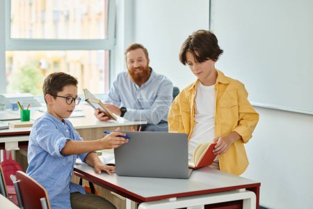 A young boy sits attentively at a laptop while a teacher observes closely in a bright and lively classroom setting.
