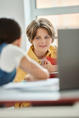 Foto de Un chico joven se sienta en un escritorio en una clase vibrante, absorto en la pantalla de su computadora portátil - Imagen libre de derechos