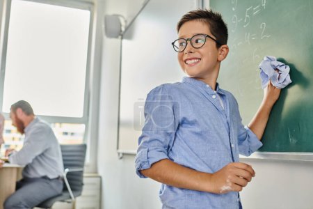 enfant se tient devant un tableau noir, souriant dans un cadre de classe dynamique.