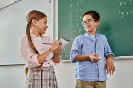 Téléchargez les photos : Un garçon et une fille debout devant un tableau noir dans une salle de classe lumineuse - en image libre de droit
